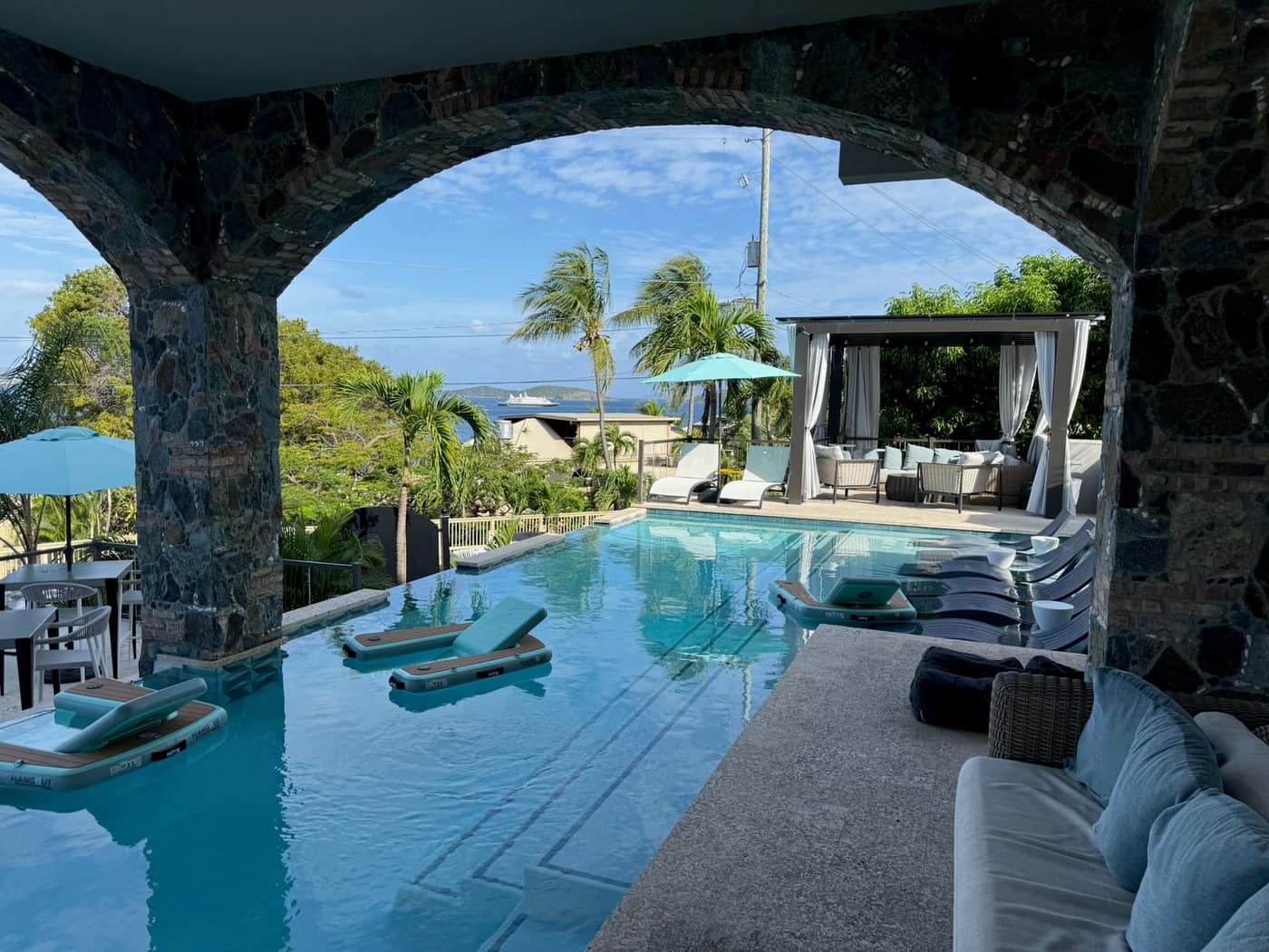 Pool deck at The Saint, St. John, at dusk with rattan pendant lights, lounge floats in the water, and the harbor beyond.