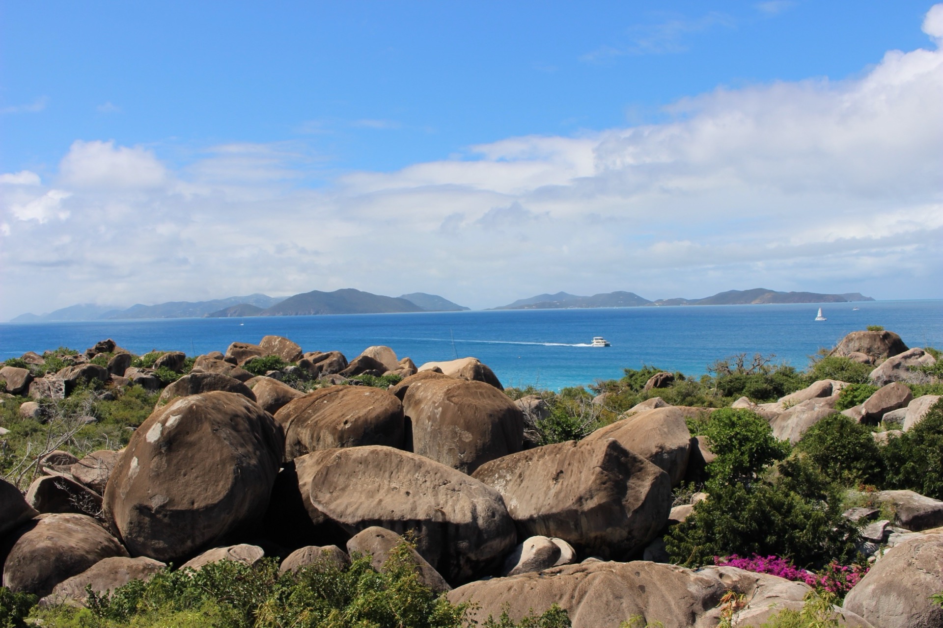 Granite boulders at The Baths on Virgin Gorda, with a powerboat passing on turquoise water and hills across the Sir Francis Drake Channel in the distance.