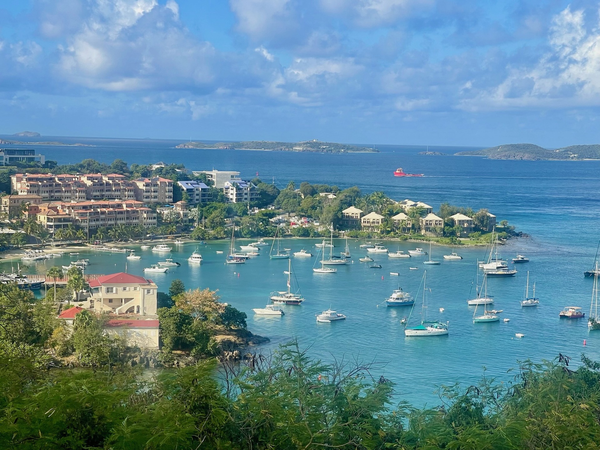 Aerial view of Cruz Bay harbor on St. John, with sailboats and powerboats moored in a turquoise anchorage, hillside villas along the shore, and a small island visible across the water.